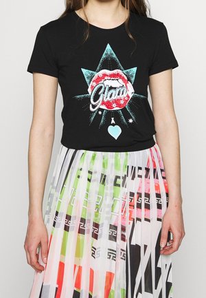 Frau trägt ein schwarzes T-Shirt mit roten Lippen und dem Text "Glam" über einem blauen Stern, kombiniert mit einem plissierten Rock in mehrfarbigem geometrischem Muster.