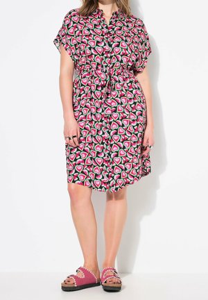 Frau trägt ein kurzärmeliges, vorne geknöpftes Kleid mit buntem Wassermelonenmuster und pinken Sandalen mit wellenförmigen Rändern, steht vor weißem Hintergrund.