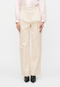 Φαρδιά παντελόνια σε ανοιχτό μπεζ χρώμα με υφή στο ύφασμα, πιέτες μπροστά, ψηλή μέση και πλαϊνές τσέπες, συνδυασμένα με μυτερά μαύρα παπούτσια.