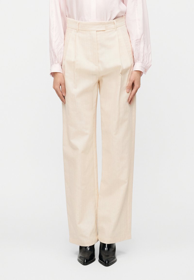 Φαρδιά παντελόνια σε ανοιχτό μπεζ χρώμα με υφή στο ύφασμα, πιέτες μπροστά, ψηλή μέση και πλαϊνές τσέπες, συνδυασμένα με μυτερά μαύρα παπούτσια.