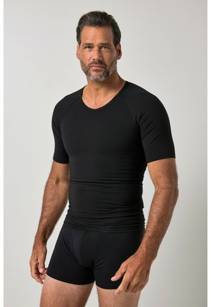Hombre con barba corta que lleva una camiseta ajustada negra de manga corta y calzoncillos tipo bóxer negros, de pie frente a un fondo liso y claro.