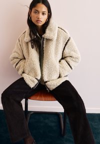 Giovane donna con lunghi capelli scuri, che indossa una giacca beige in shearling e pantaloni neri, seduta su una sedia di legno contro un muro chiaro.