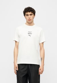 Νεαρός άνδρας με σγουρά μαλλιά που φοράει λευκό T-shirt Puma και της ομάδας Formula One της Aramco και μαύρο παντελόνι, στέκεται μπροστά σε απλό φόντο.