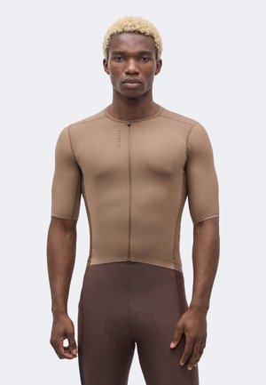 Junger Mann mit blondem lockigem Haar, der ein eng anliegendes braunes kurzärmeliges Radtrikot und passende gepolsterte Shorts trägt, stehend und nach vorne schauend.