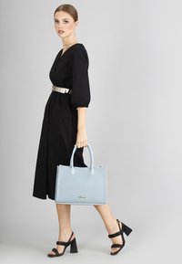 Borsa tote in pelle blu chiaro con doppie maniglie e cucitura a contrasto, abbinata a un vestito nero con maniche lunghe e una cintura metallica.