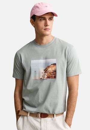 Jonge man draagt een grijs T-shirt met een afbeelding van een kustklif en de tekst "GANT NANTUCKET", een roze pet, een beige riem en lichtgekleurde broek.