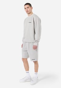 Sudadera gris claro y pantalones cortos a juego, hechos de una tela suave. Presenta puños acanalados y un logo negro en el pecho y el dobladillo inferior. Zapatillas deportivas blancas.