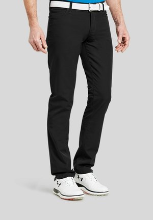 Man draagt een zwarte broek, witte riem, blauw overhemd en witte Under Armour golfschoenen, staand op een grijze achtergrond.