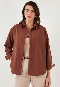 Brown button-up shirt made of soft fabric, featuring a classic collar, long sleeves rolled at cuffs, and a relaxed fit.