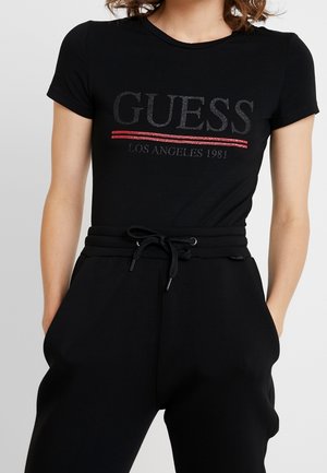 Femme portant un t-shirt noir près du corps avec le texte « GUESS Los Angeles 1981 » souligné en rouge, associé à un pantalon noir à cordon.