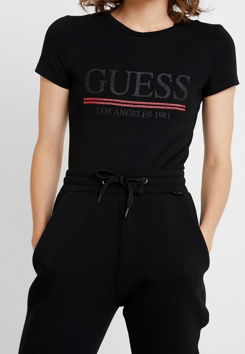 Femme portant un t-shirt noir près du corps avec le texte « GUESS Los Angeles 1981 » souligné en rouge, associé à un pantalon noir à cordon.