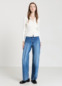 Weißer gerippter Cardigan mit Knopfverschluss, kombiniert mit blauen weiten Jeans, die auffällige Nähte und eine lockere Passform aufweisen. Schwarze Schuhe.