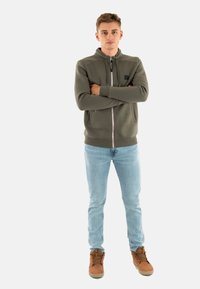 Felpa verde con zip e collo alto, dettagli grigi e una toppa con logo. Abbinata a jeans blu chiaro e stivali marroni con lacci.