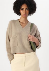 Maglione beige corto con scollo a V, texture a coste e due tasche frontali. Abbinato a pantaloni giallo chiaro e una borsa a spalla verde.