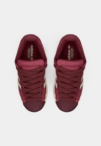 Ζευγάρι μπορντό αθλητικά παπούτσια Adidas με λευκές ρίγες και προστατευτικά στα δάχτυλα σε σχήμα κελύφους, σε λευκό φόντο, φωτογραφημένα από πάνω.