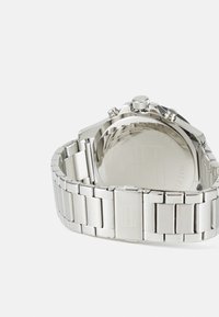 Silberne Metalluhr mit einem polierten Band und gebürsteten Gliedern. Sie verfügt über ein rundes Gehäuse, eine strukturierte Krone und ein graviertes Logo am Verschluss.