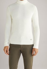 Mies, joka pukeutuu valkoiseen ribattuun poolopaitaan ja oliivinvihreisiin housuihin, seisoo yksinkertaista taustaa vasten.