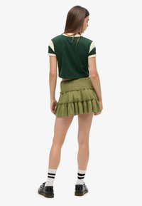 Grünes, bauchfreies Shirt mit cremefarbenen Ärmeln, kombiniert mit einem Stufensrock in Olivgrün. Accessoires sind schwarze Schuhe und gestreifte Socken.