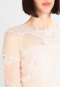 Femme portant une robe en dentelle rose clair avec des motifs floraux et un tissu transparent, montrant des détails de l'épaule à la partie supérieure de la poitrine.