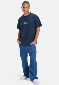 Marineblå bomull T-skjorte med grønn Quiksilver-logo, kombinert med blå denimjeans. T-skjorten har en avslappet passform og korte ermer.