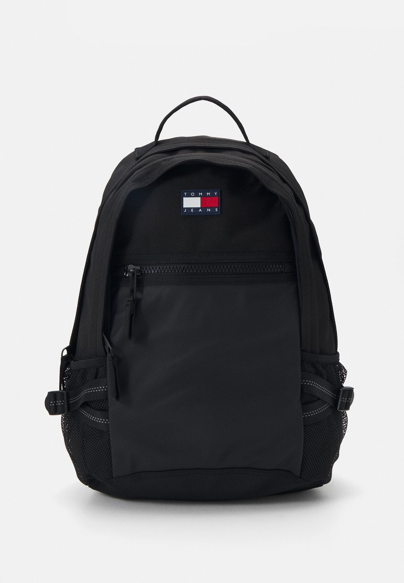 Mochila negra con un acabado de tela suave, que cuenta con un bolsillo frontal con cremallera, bolsillos laterales de malla y un parche con el logotipo en la parte superior.