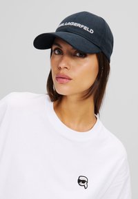 Marineblaue Baseballmütze mit gebogenem Schirm und gesticktem Schriftzug "KARL LAGERFELD". Kombiniert mit einem weißen T-Shirt, das ein kleines grafisches Logo trägt.