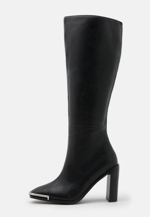 Bota negra texturizada hasta la rodilla con punta puntiaguda, detalle metálico plateado en la punta y tacón grueso sobre fondo blanco.