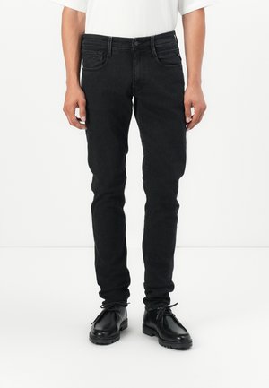 Homme portant un jean noir slim et des chaussures noires à lacets, debout devant un fond blanc uni.