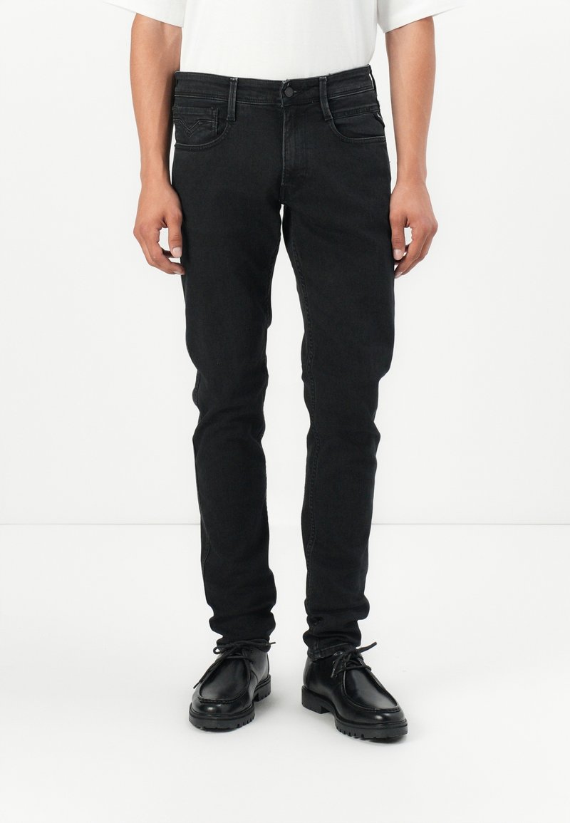 Homme portant un jean noir slim et des chaussures noires à lacets, debout devant un fond blanc uni.