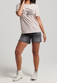 Světle šedé bavlněné tričko s grafikou "Superdry", doplněné o tmavé džíny šortky. Model má na sobě bílé vysoké tenisky. Jednoduchý design.