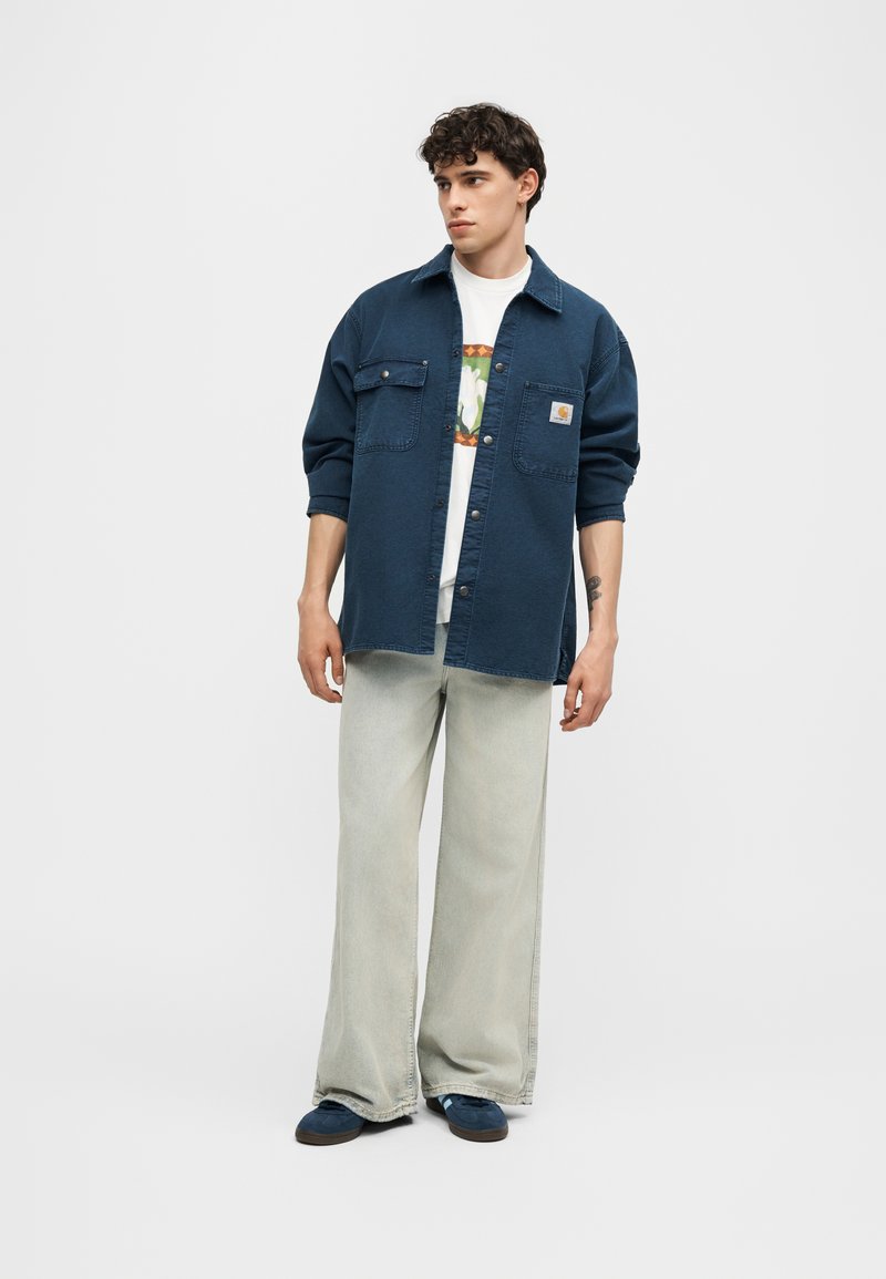 Junger Mann trägt ein marineblaues Overshirt, ein weißes grafisches T-Shirt, weite hellgraue Jeans und blaue Sneakers und steht vor einem einfarbigen Hintergrund.