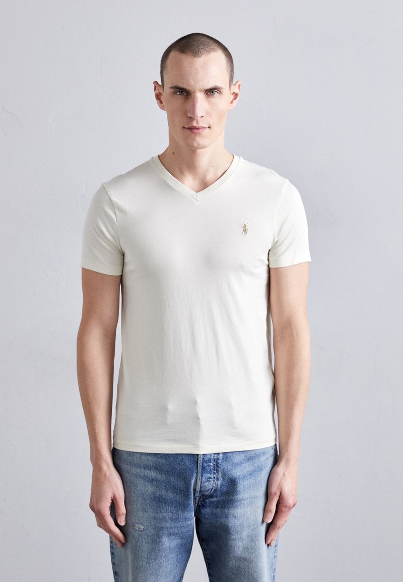 T-shirt beige clair à col en V en coton doux, avec des manches courtes et petit logo sur la poitrine, associé à un jean en denim bleu.