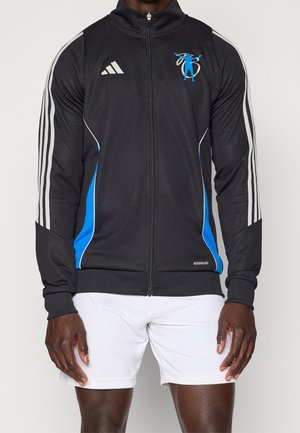 Männliches Model trägt schwarze Adidas Zip-Up-Jacke mit weißen Streifen und blauen Akzenten, kombiniert mit weißen Sportshorts, stehend vor einfarbigem Hintergrund.