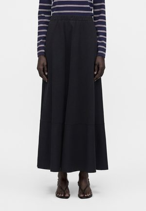 Femme portant une longue jupe noire, un haut à manches longues rayé bleu marine et blanc, et des sandales noires à brides, se tenant devant un fond uni.