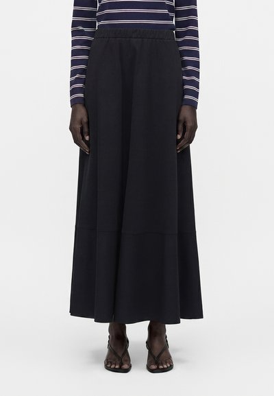 Femme portant une longue jupe noire, un haut à manches longues rayé bleu marine et blanc, et des sandales noires à brides, se tenant devant un fond uni.