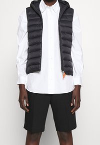 Chaleco tipo puff negro con capucha, cierre de cremallera y tirador de cremallera naranja, usado sobre una camisa blanca de botón, combinado con pantalones cortos negros.