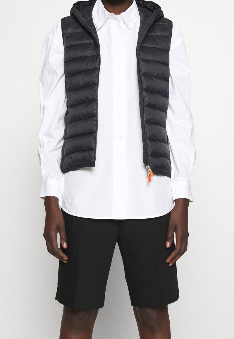 Chaleco tipo puff negro con capucha, cierre de cremallera y tirador de cremallera naranja, usado sobre una camisa blanca de botón, combinado con pantalones cortos negros.