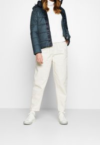 Ett vadderat mörkblått jacka med huva, vit texturerad tröja, krämfärgade byxor och vita sneakers med texturerade sulor, står mot en ljus bakgrund.