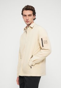 Νέος άνδρας που φοράει μπεζ ζακέτα με φερμουάρ και διακοσμητικό ταμπελάκι BOSS στην τσέπη του αριστερού μανικιού, μαύρο παντελόνι, στέκεται με το ένα χέρι στην τσέπη.