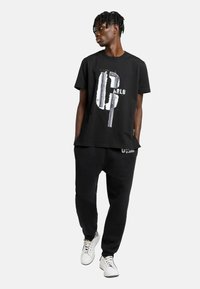 Ung mand med kort snoet hår iført sort grafisk T-shirt, sorte joggers og hvide sneakers, stående med hænderne i lommerne og ser til venstre.