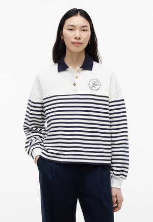 Jonge vrouw die een wit en marineblauw gestreepte lange mouwen polo met marineblauwe kraag en donkere broek draagt, staand met één hand in de zak.
