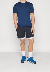 Muž v modrém tričku Under Armour, černých šortkách s bílými kompresními šortkami, bílých sockách a modrých teniskách stojící na bílém pozadí.