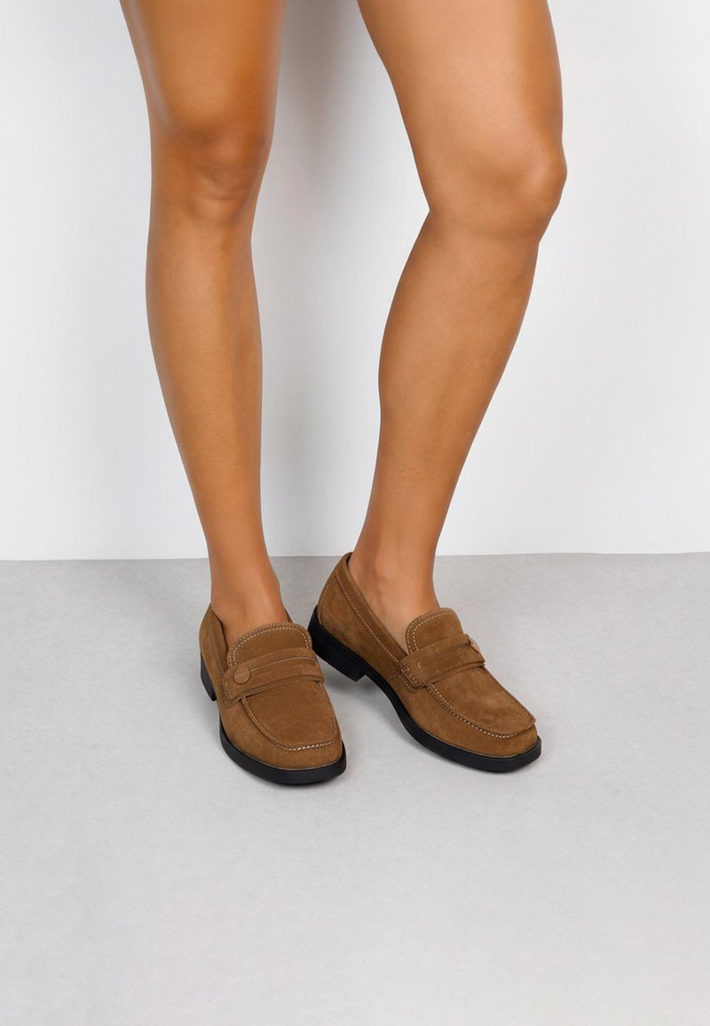 Mocasines de ante marrón con un tacón bajo, que presentan un diseño de delantal cosido y un acento de botón decorativo, combinados con piernas al aire.