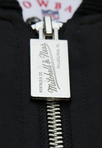 Schwarzer Stoffreißverschluss mit einem rechteckigen silbernen Ziehring, auf dem "Mitchell & Ness" eingraviert ist. Glatte Textur mit metallischen Hardware-Details.