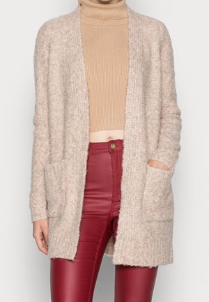 Cardigan beige duveteux avec poches devant porté sur un col roulé court côtelé couleur fauve et un pantalon en cuir synthétique rouge taille haute.