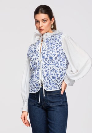 Vrouw met een witte blouse met ruches aan de kraag en een blauw gebloesd patroon gilet met een hartvormige zak, gecombineerd met een donkere spijkerbroek.