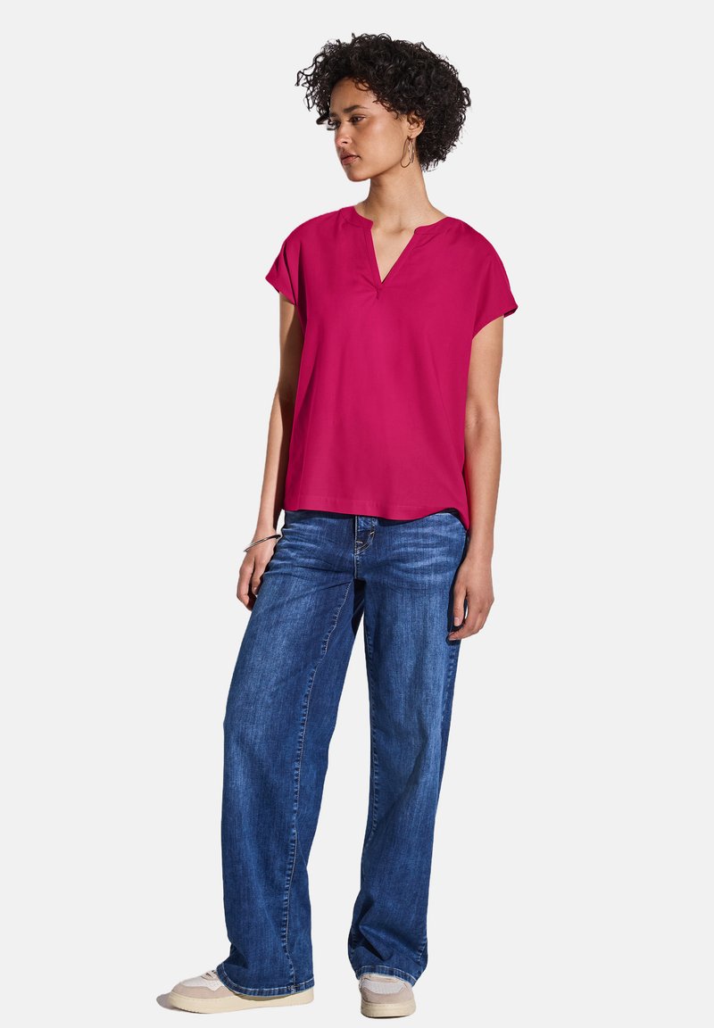 Top magenta a maniche corte con scollo a V, abbinato a jeans blu a gamba larga. Tessuto liscio e vestibilità rilassata.
