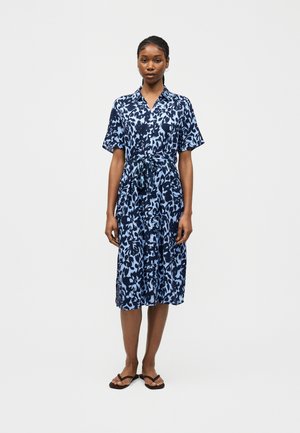 Kvinde stående iført en blå og navy knap-op kjole med blomsterprint, korte ærmer og sorte sandaler på en ensfarvet hvid baggrund.