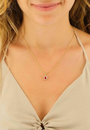Collier chaîne en or avec un pendentif rubis ovale, entouré de petites pierres transparentes, porté sur un haut beige clair.