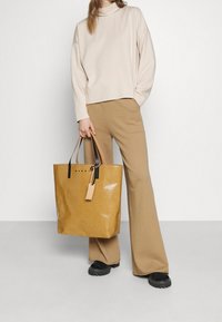 Tote bag marron à finitions brillantes, avec des poignées en cuir et des accents noirs. Le mannequin porte un haut beige et un pantalon beige à pattes d'éléphant.